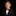 A young boy with brown hair wearing a black suit jacket, white dress shirt, and a deep red textured bow tie with a matching red pocket square, set against a solid black background.
