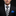 Man wearing a black suit with a white shirt, black and gray striped tie, and blue pocket square on a black background.