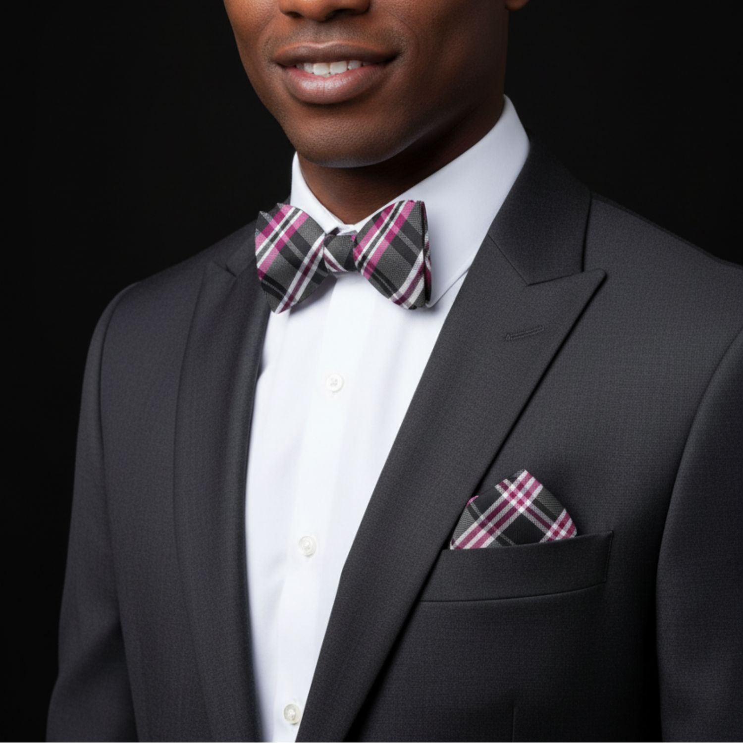 Man wearing a black suit with a plaid bow tie and pocket square on a black background
