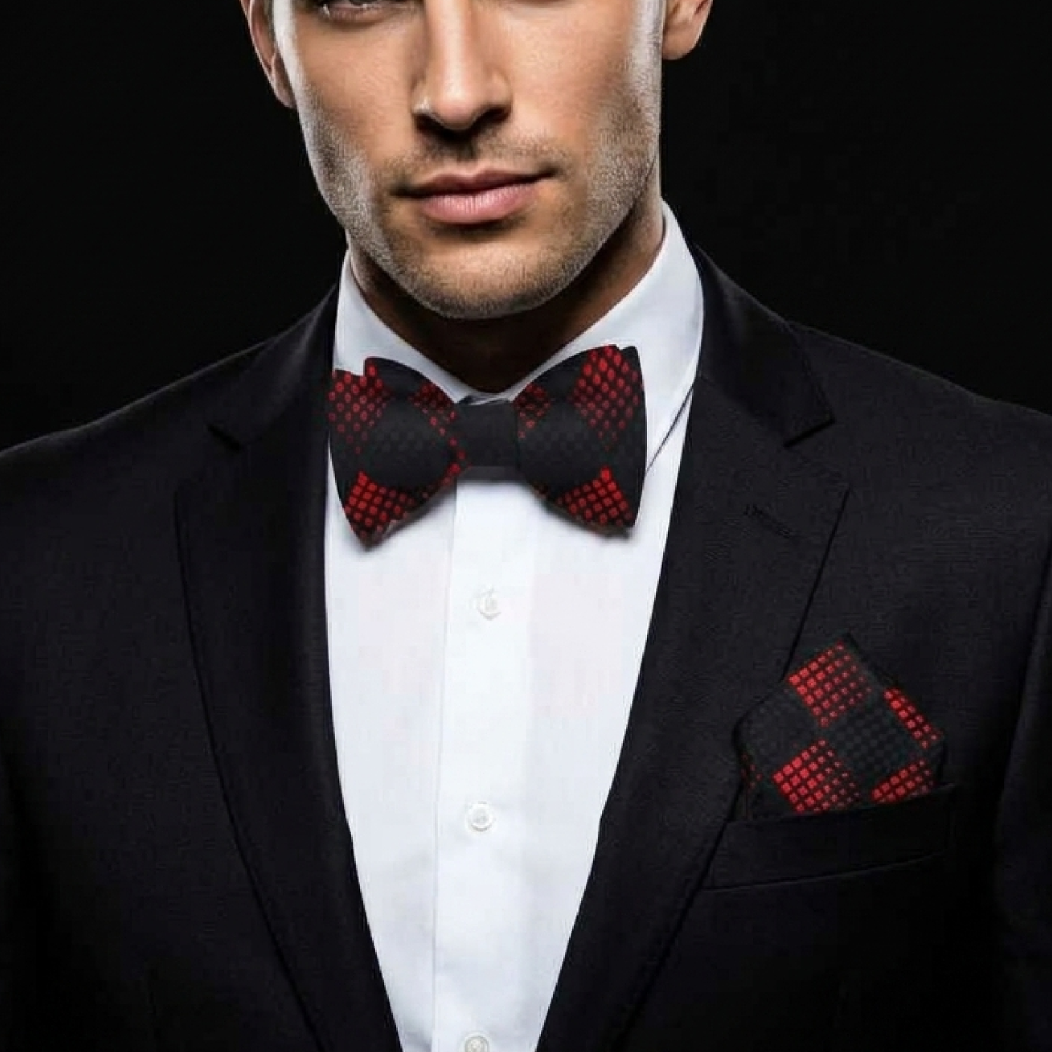 Man wearing a black suit with a red and black checkered bow tie and pocket square against a black background