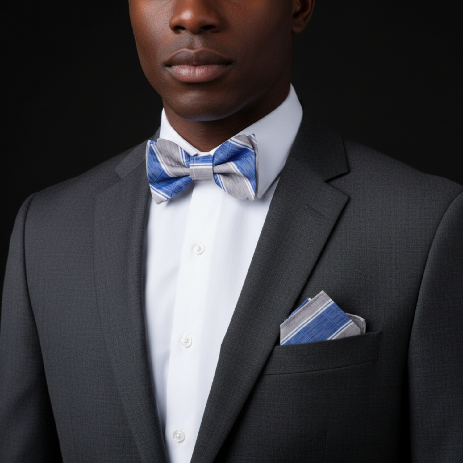 Man wearing a gray suit with a blue and gray striped bow tie and pocket square against a black background