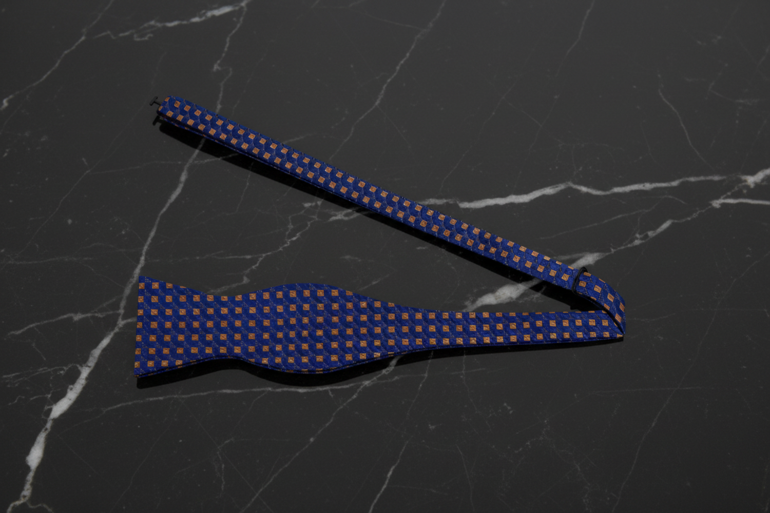 A dark blue bow tie with subtle brown checks laid out on a wooden surface, surrounded by green leaves.