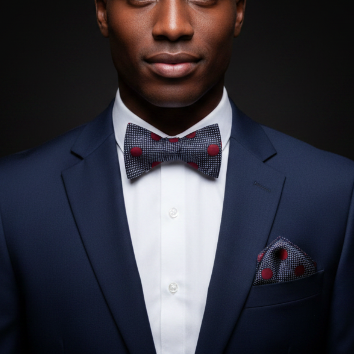 Man wearing a navy suit with a patterned bow tie and pocket square against a dark background