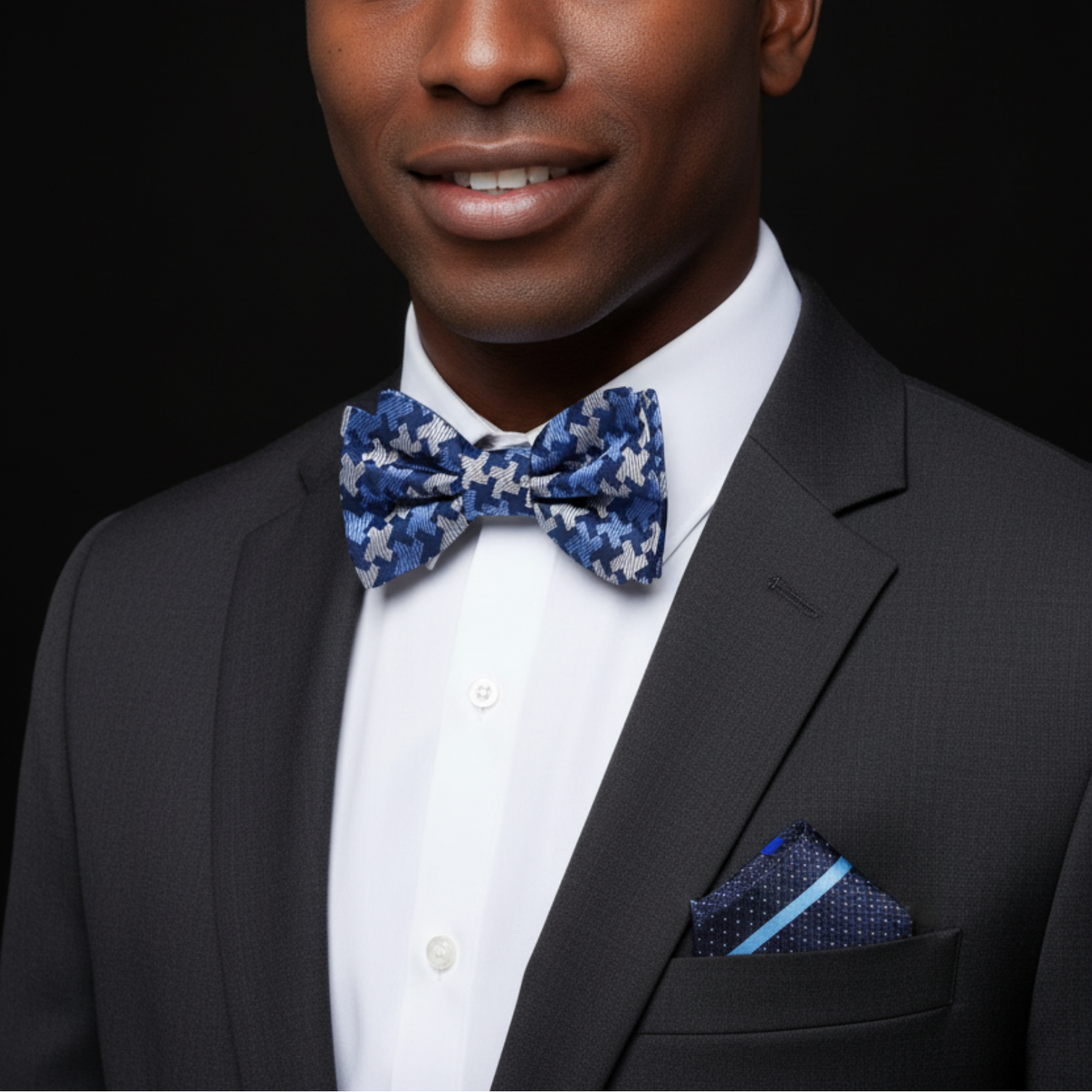 Man wearing a black suit with a blue patterned bow tie and pocket square on a black background