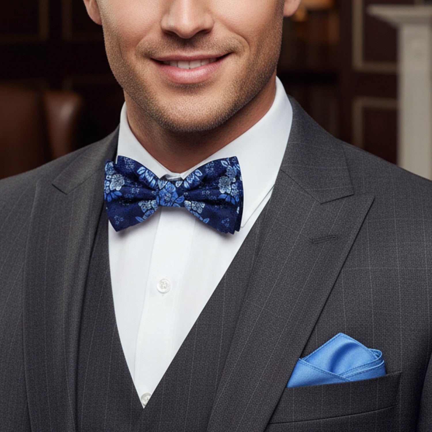 Man wearing a gray suit with a blue bow tie and light blue pocket square indoors.