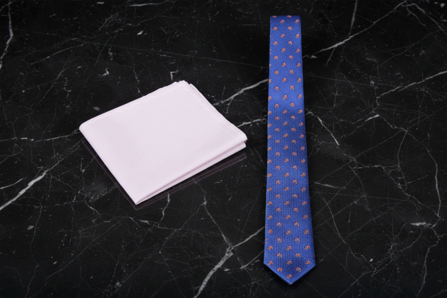 Blue patterned thin tie and pinkish white pocket square on a black marble surface