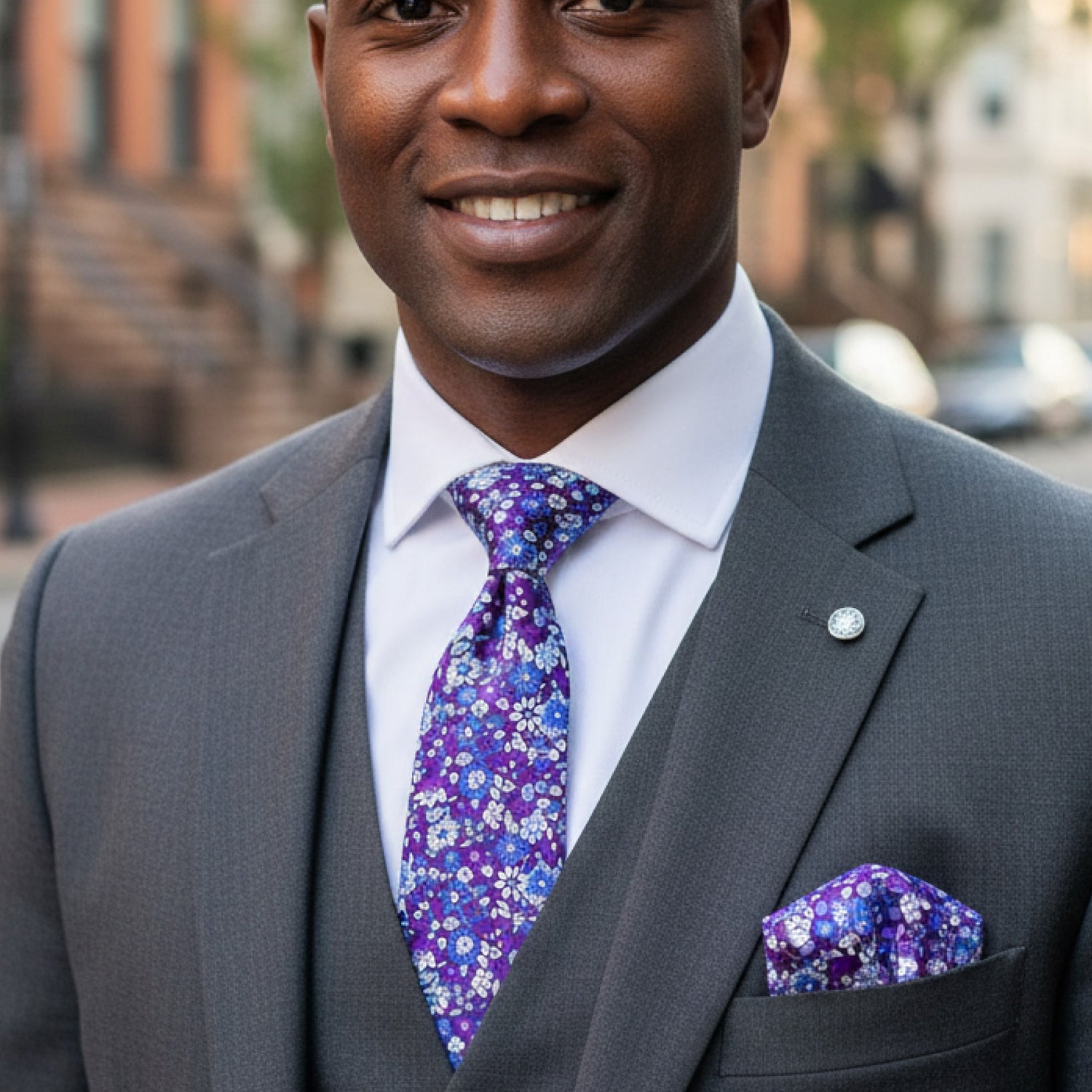 Man wearing a gray suit with a purple floral tie and pocket square, standing outdoors.