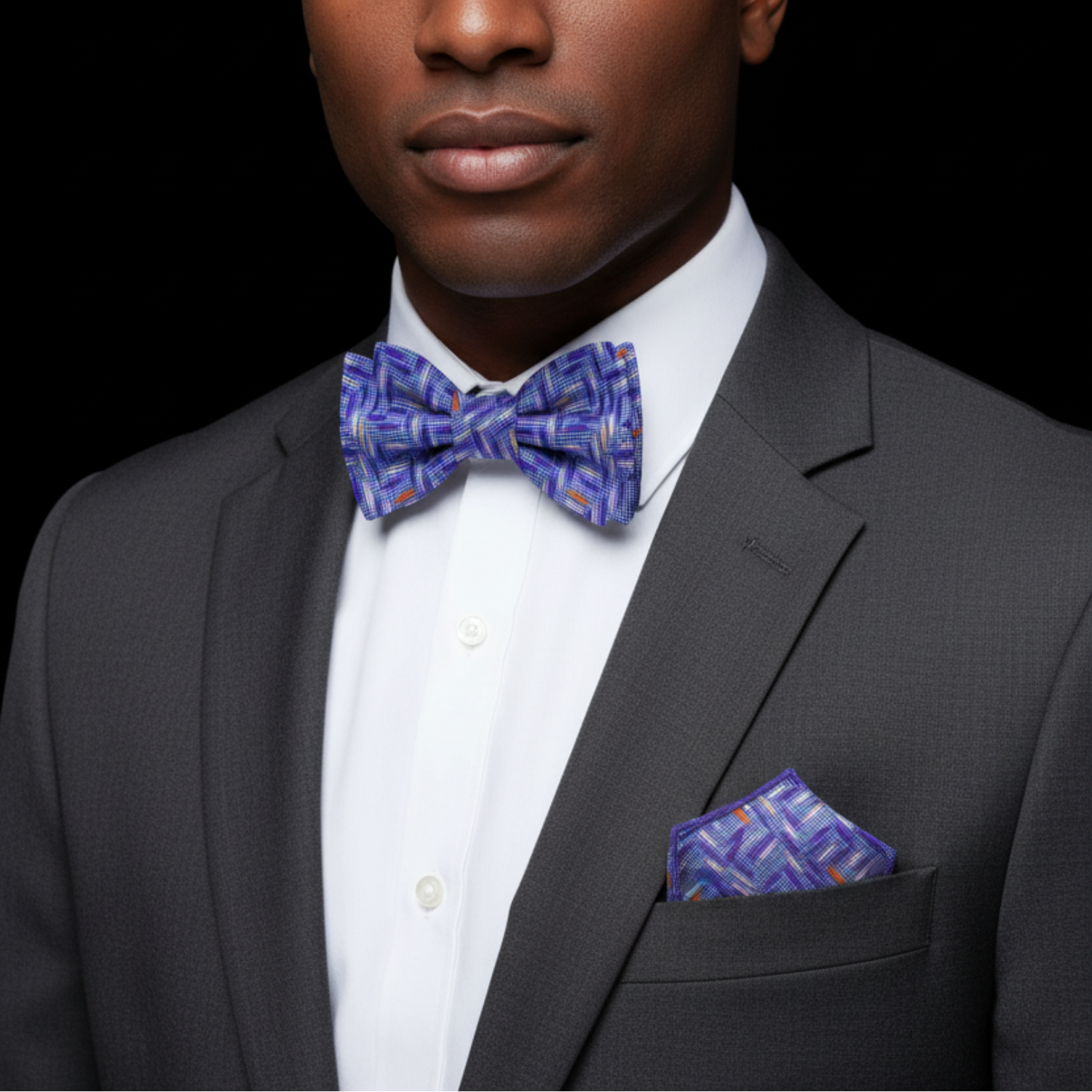 Man wearing a gray suit with a blue patterned bow tie and pocket square against a black background