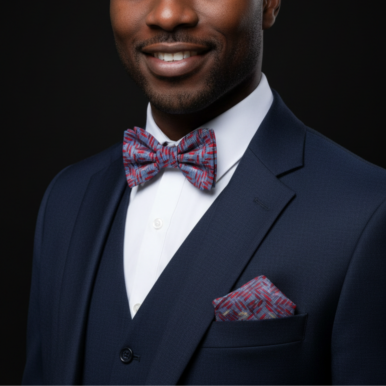 Man wearing a navy suit with a red and blue patterned bow tie and pocket square on a black background