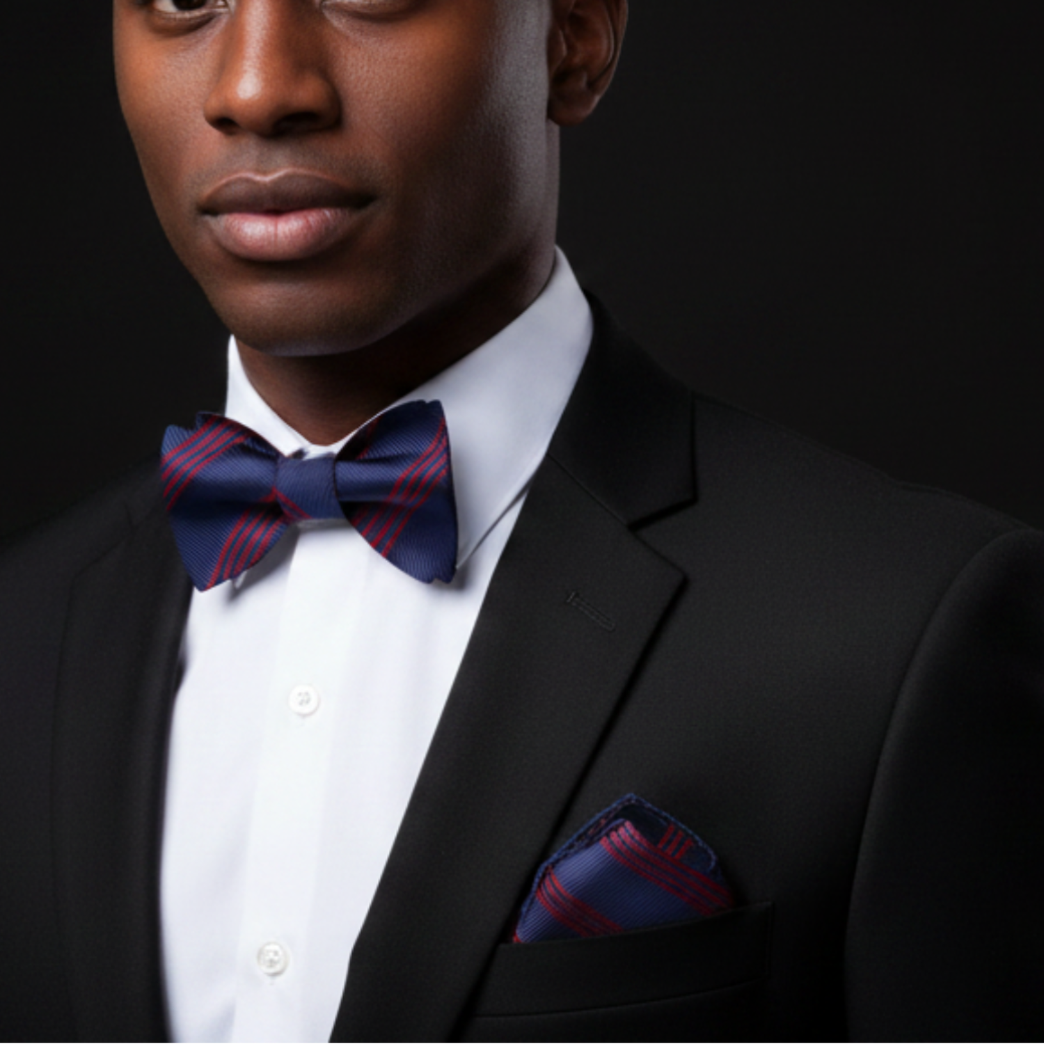 Man wearing a black suit with a white shirt and red plaid bow tie and pocket square on a dark background