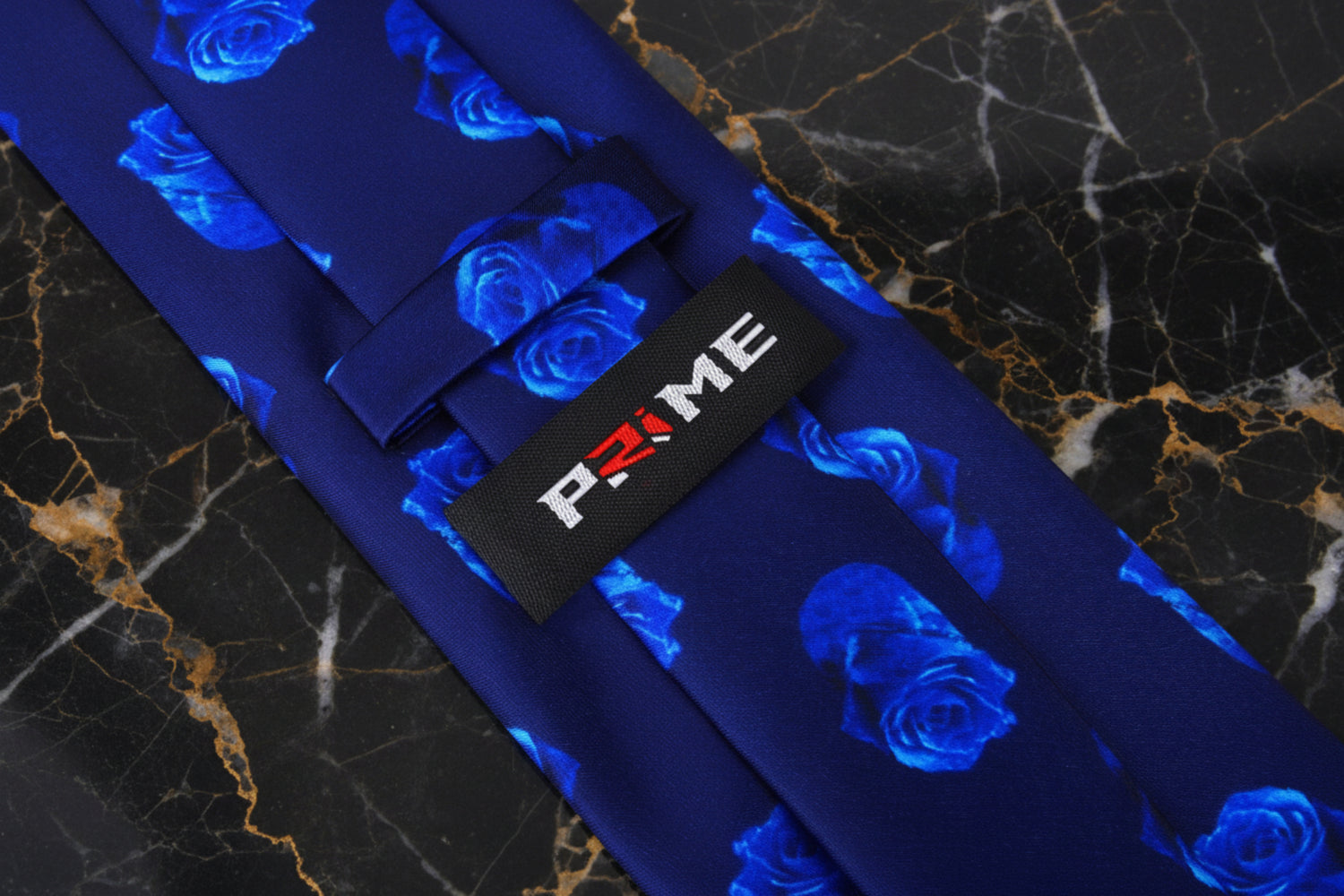 A close-up of a necktie with a dark blue satin base, a repeating pattern of vibrant blue roses, and a black fabric brand tag that reads 'P.RIME'.