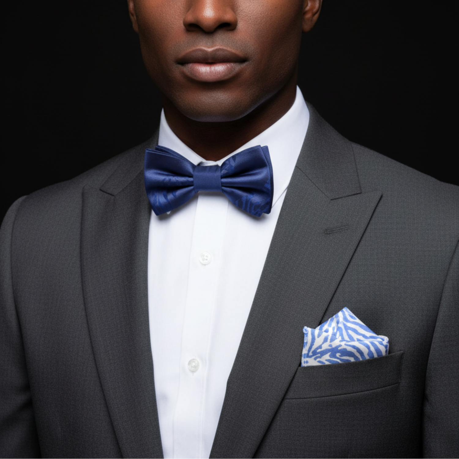 Man wearing a gray suit with a blue bow tie and patterned pocket square on a black background