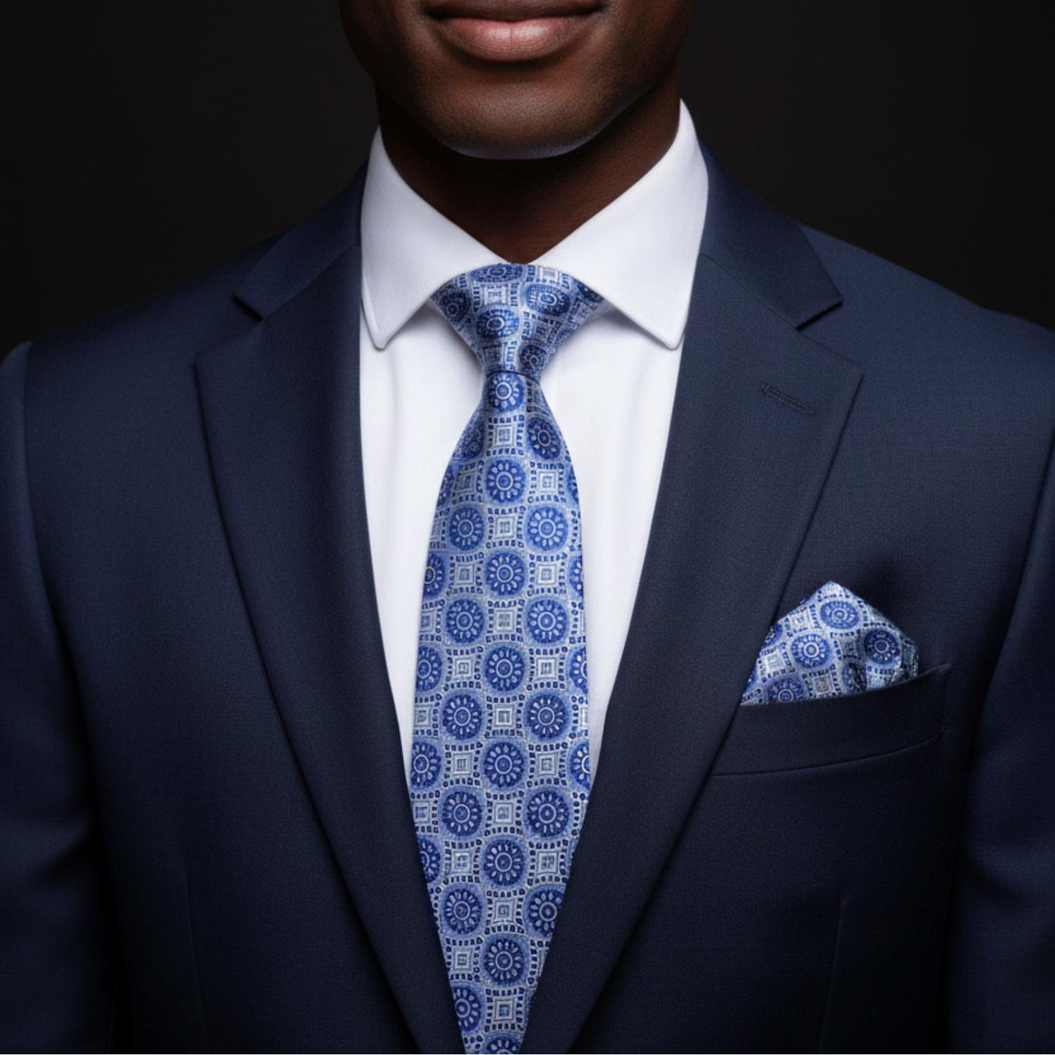 Man wearing a navy suit with a blue patterned tie and pocket square on a black background