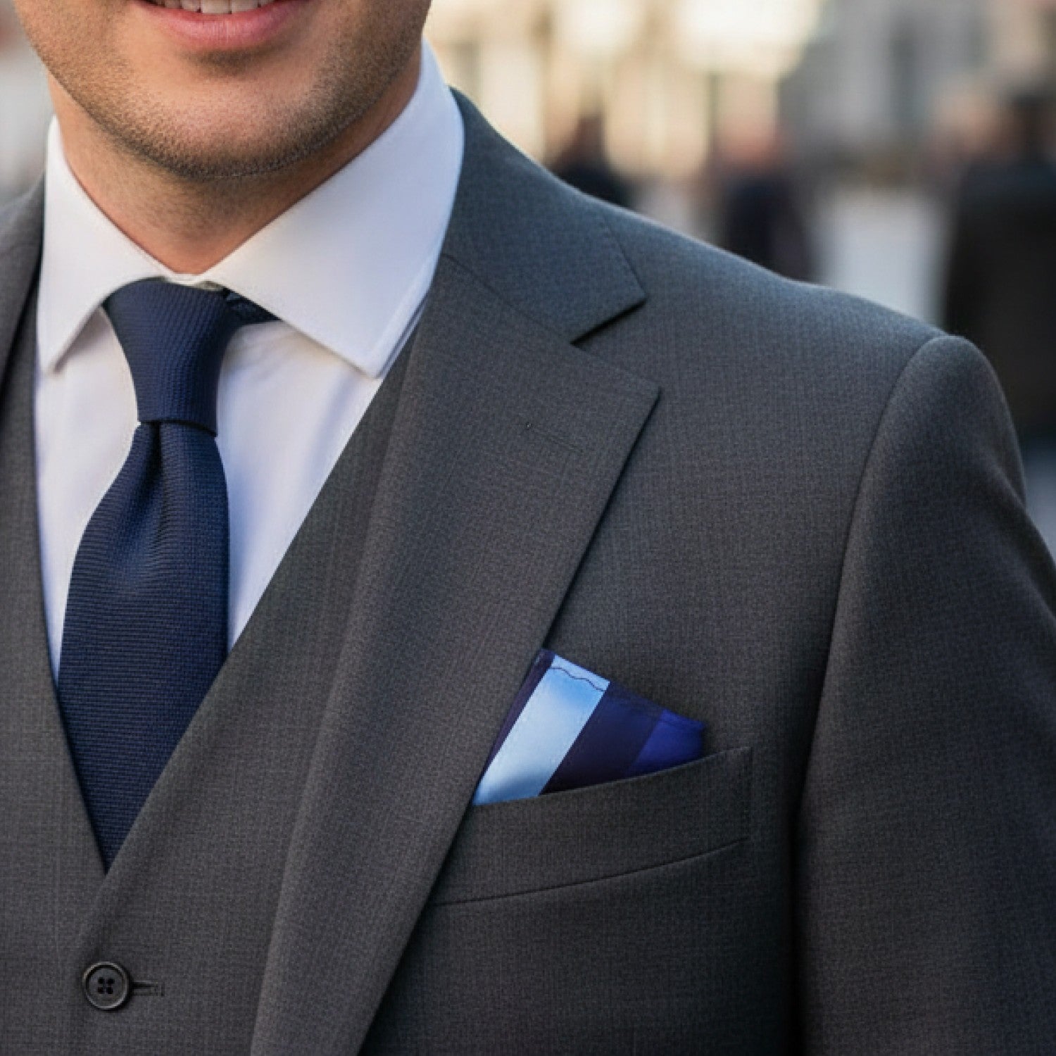 Man wearing a gray suit with a blue tie and pocket square, blurred background