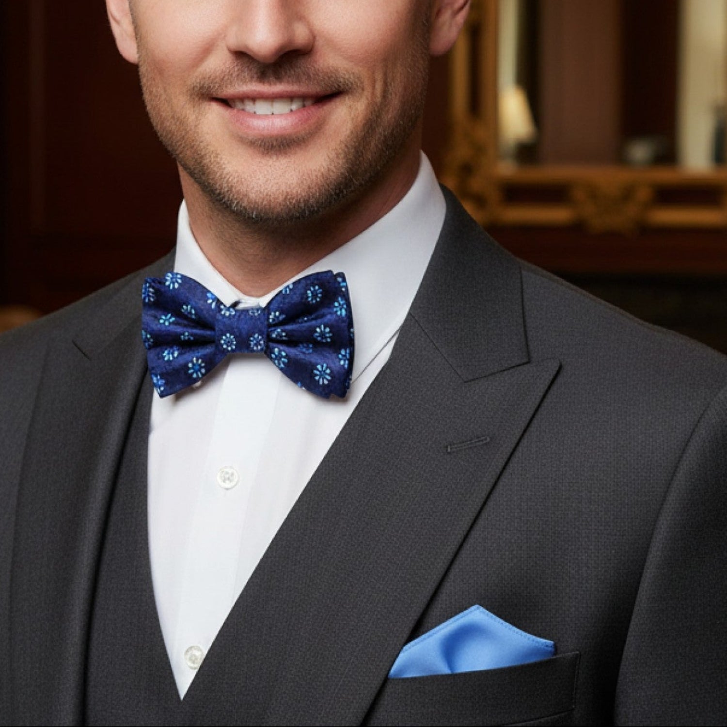 Man wearing a dark suit with a blue bow tie and pocket square in an elegant room.