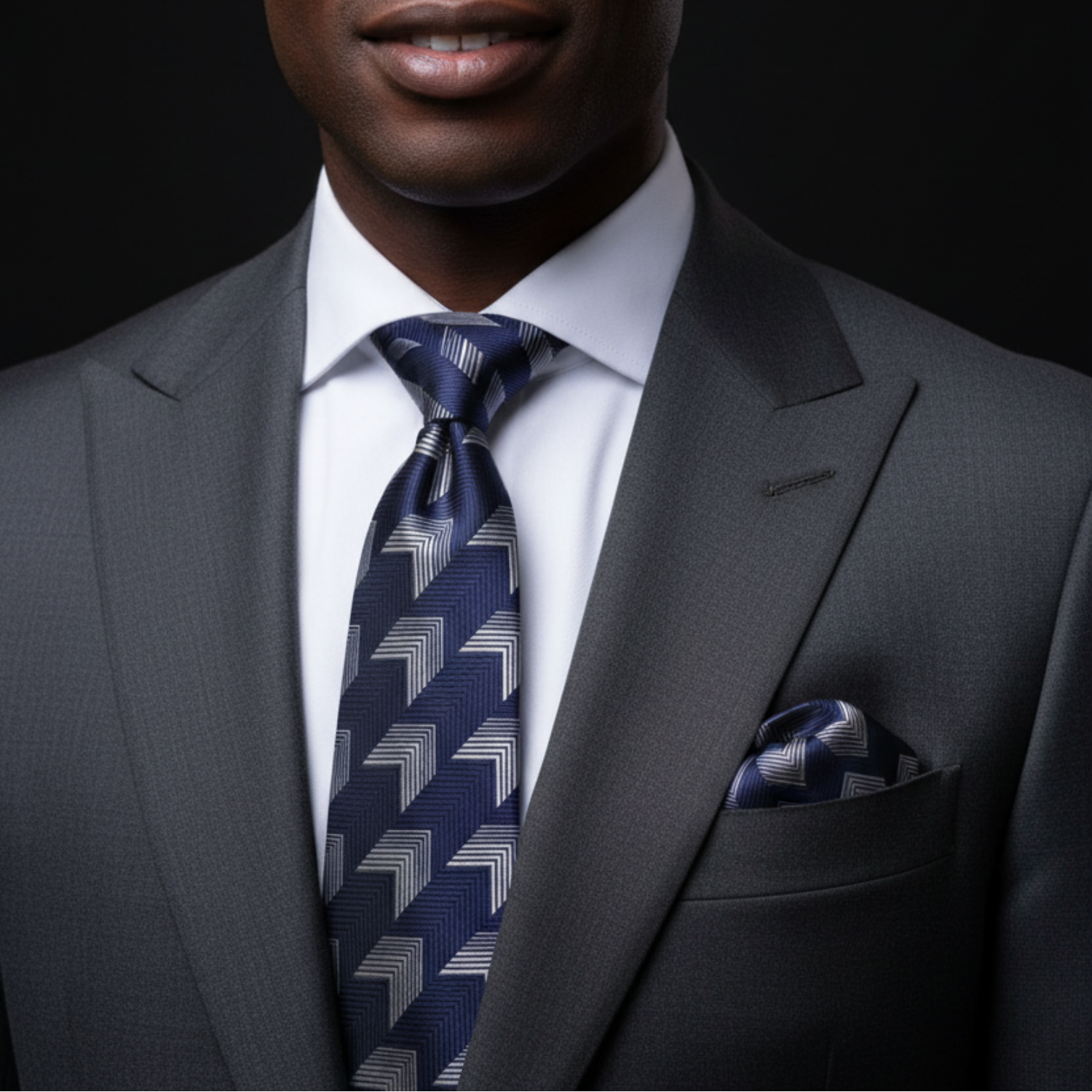 A close-up of a man wearing a dark gray suit, white dress shirt, and a navy blue patterned tie with a matching pocket square.