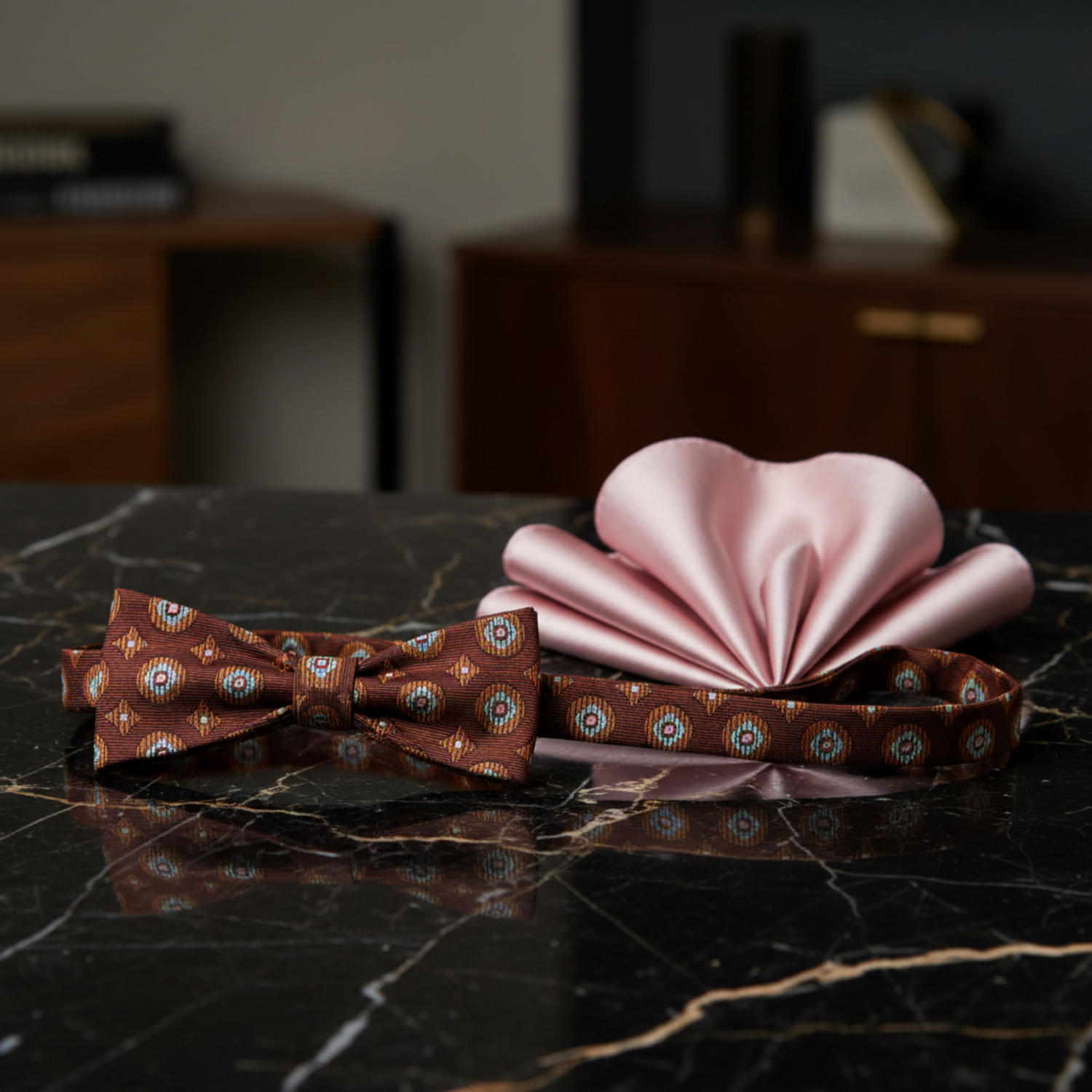 A men's formal accessory set featuring a brown patterned bow tie, a matching brown patterned necktie clip, and a light pink satin pocket square, arranged on a black marble surface.