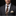 A close-up of a man wearing a dark gray suit, white dress shirt, brown patterned tie, and a cream pocket square with small red dots, set against a solid black background.