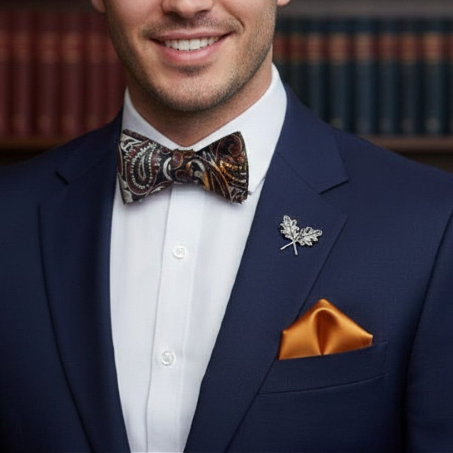 Man in a blue suit with a patterned bow tie and orange pocket square, standing in front of bookshelves.