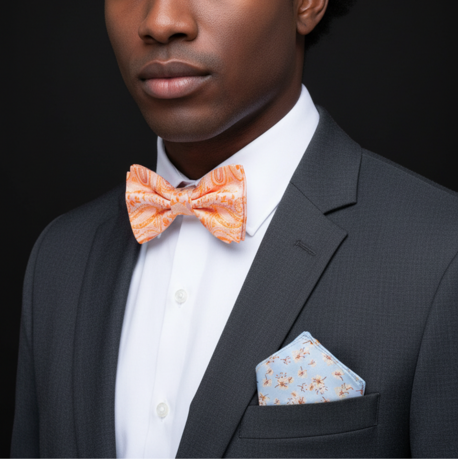 Man wearing a gray suit with an orange bow tie and light blue pocket square against a black background