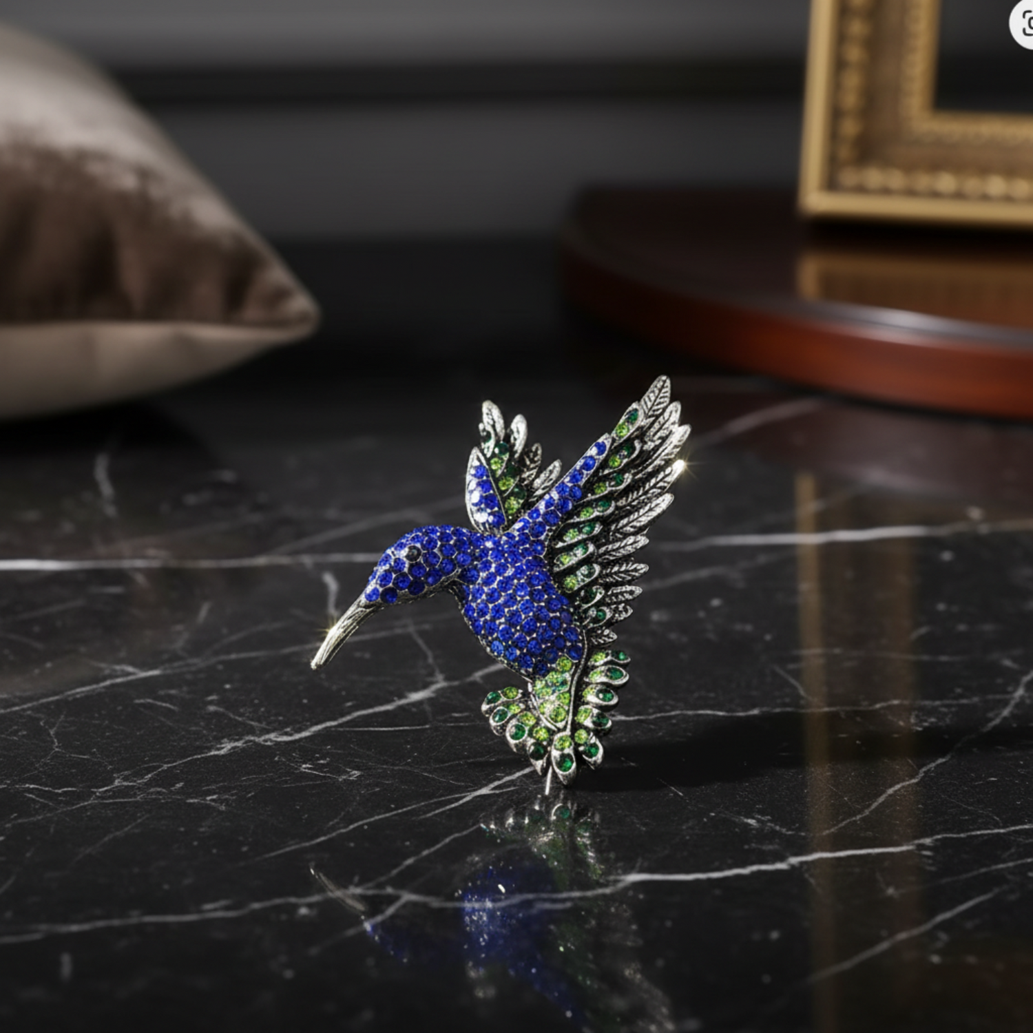 Decorative hummingbird brooch with blue and green gemstones held in a hand against a neutral background.