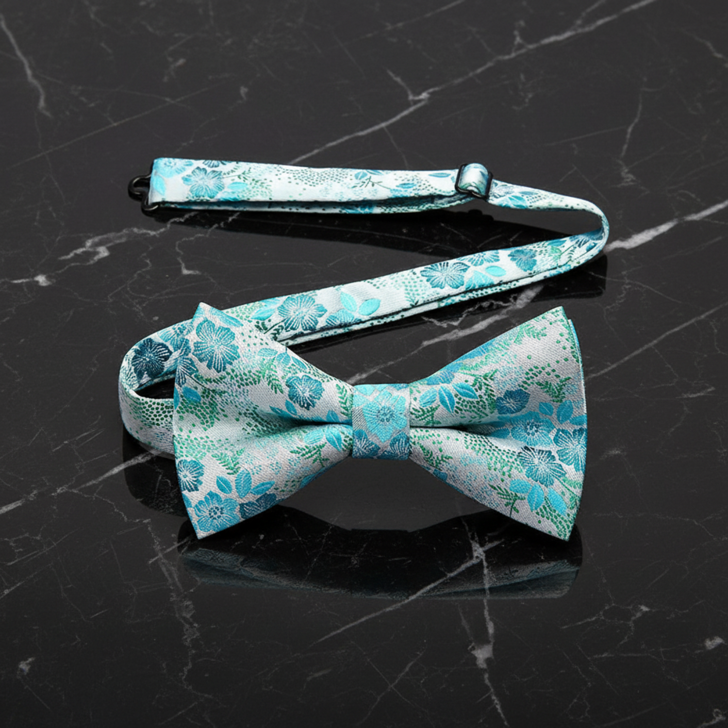 A floral bow tie with a combination of light teal, green, and blue colors laid out on a flat surface with a pink rose in the background.