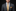 Man wearing a gray suit with a gold tie and pocket square on a black background