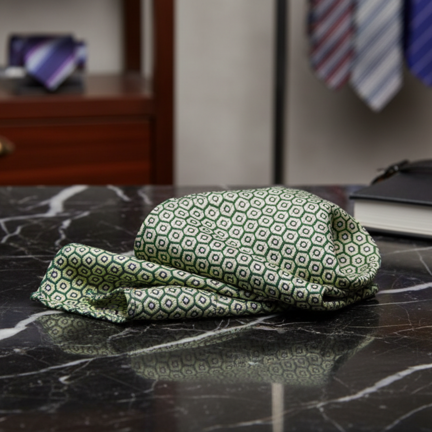 Folded green patterned handkerchief on a marble surface with ties hanging in the background