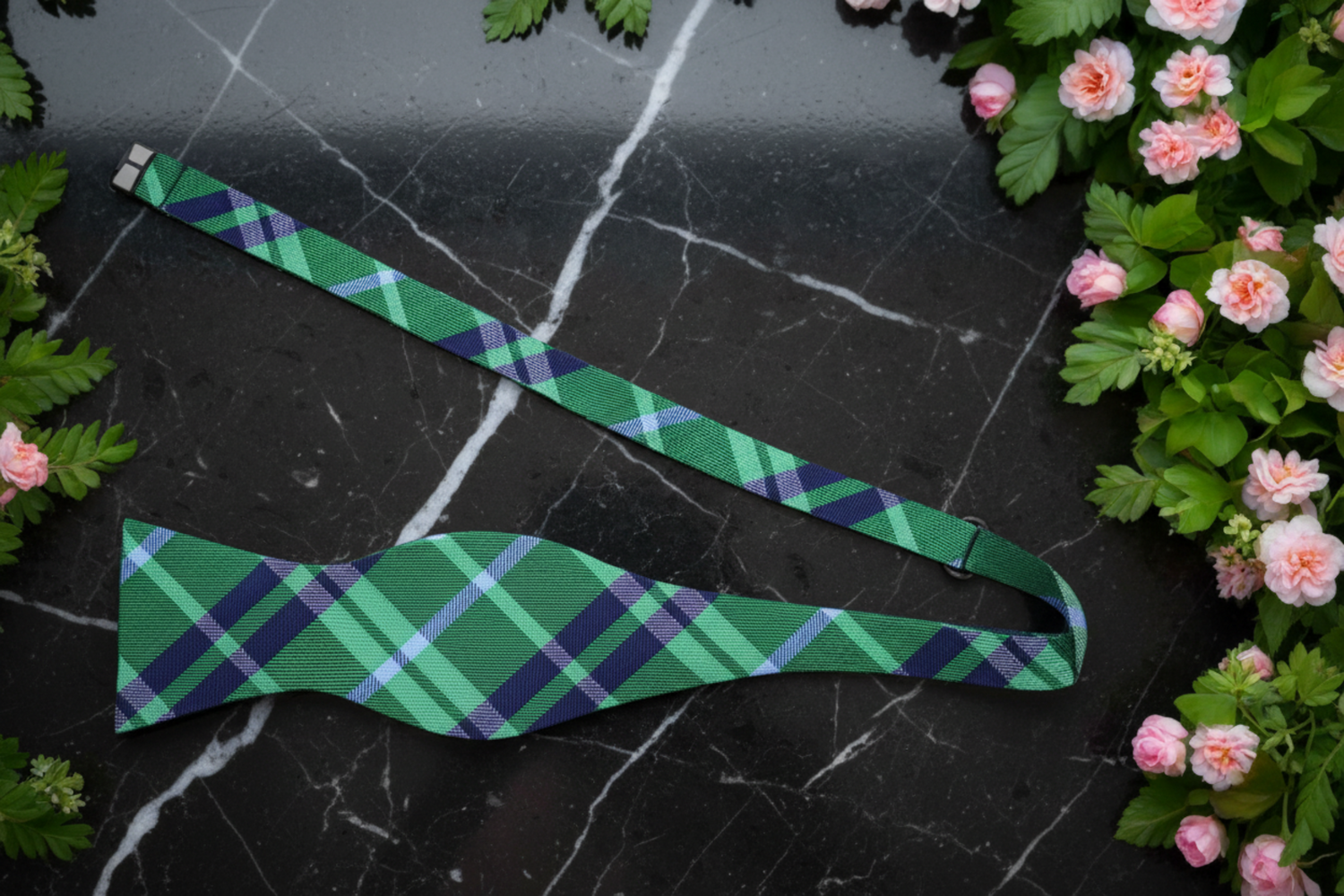Green plaid bow tie on a stone surface with flowers and leaves.