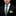 Man wearing a black suit with a green and gray plaid tie and a patterned pocket square on a black background