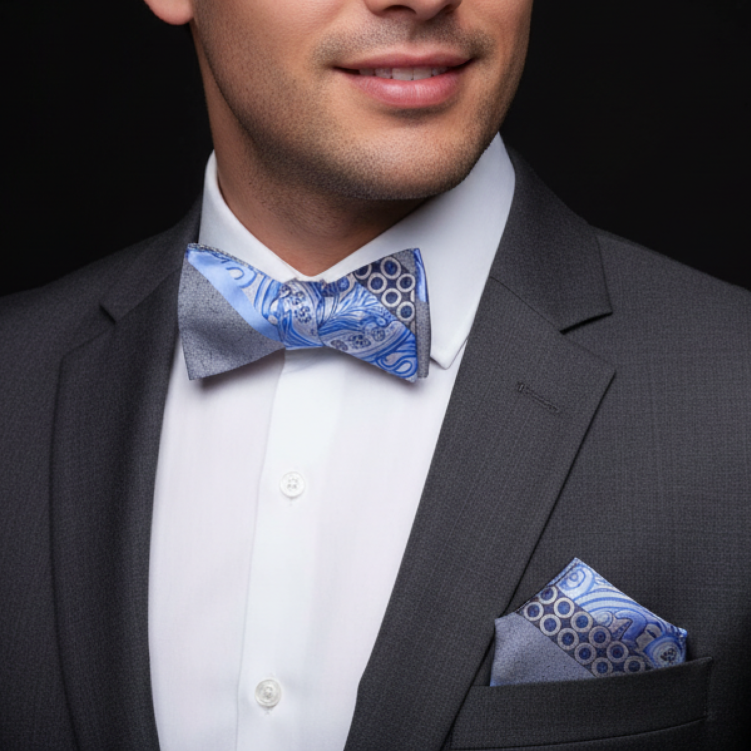 Man wearing a gray suit with a blue and silver bow tie and pocket square against a black background