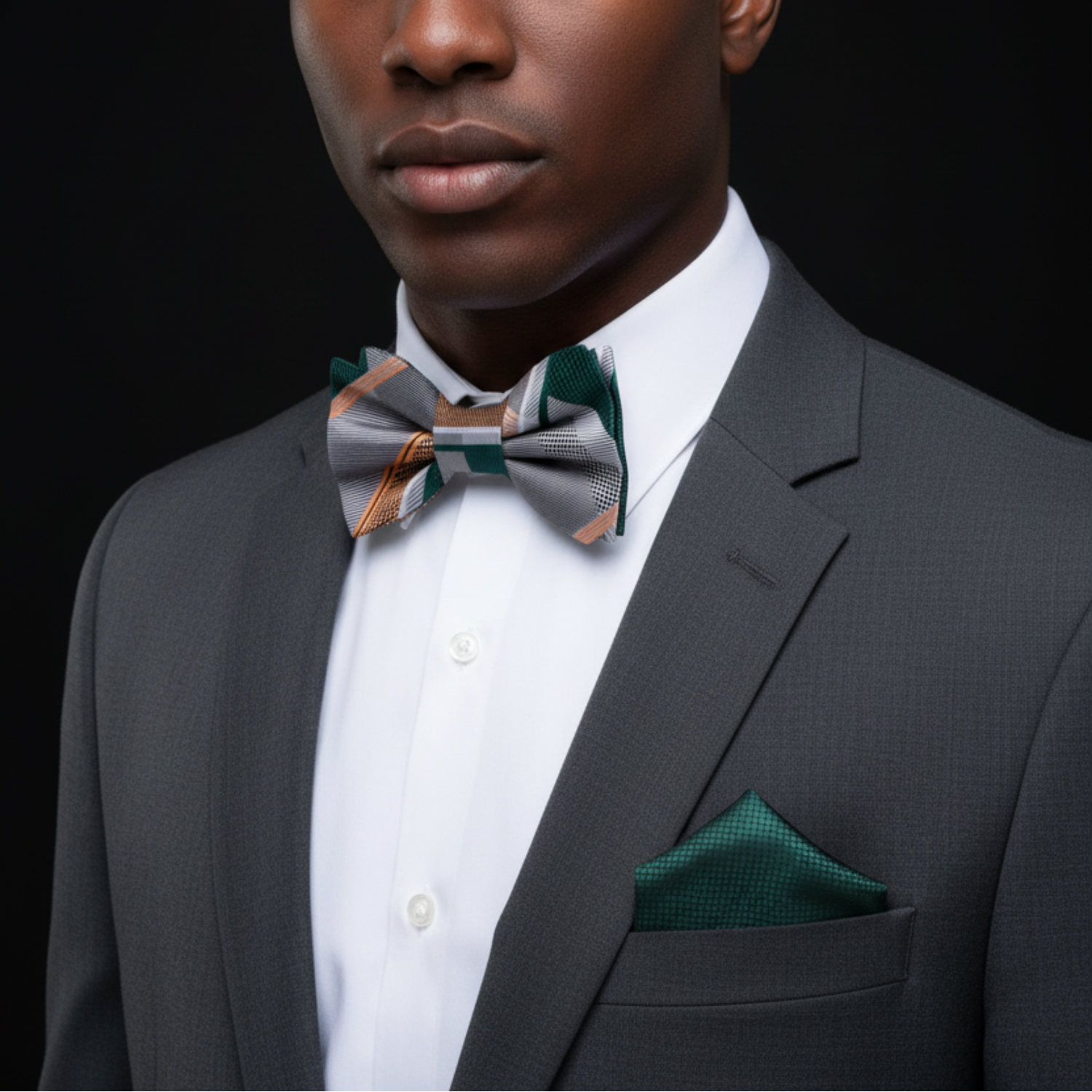 Man wearing a gray suit with a patterned bow tie and green pocket square on a black background