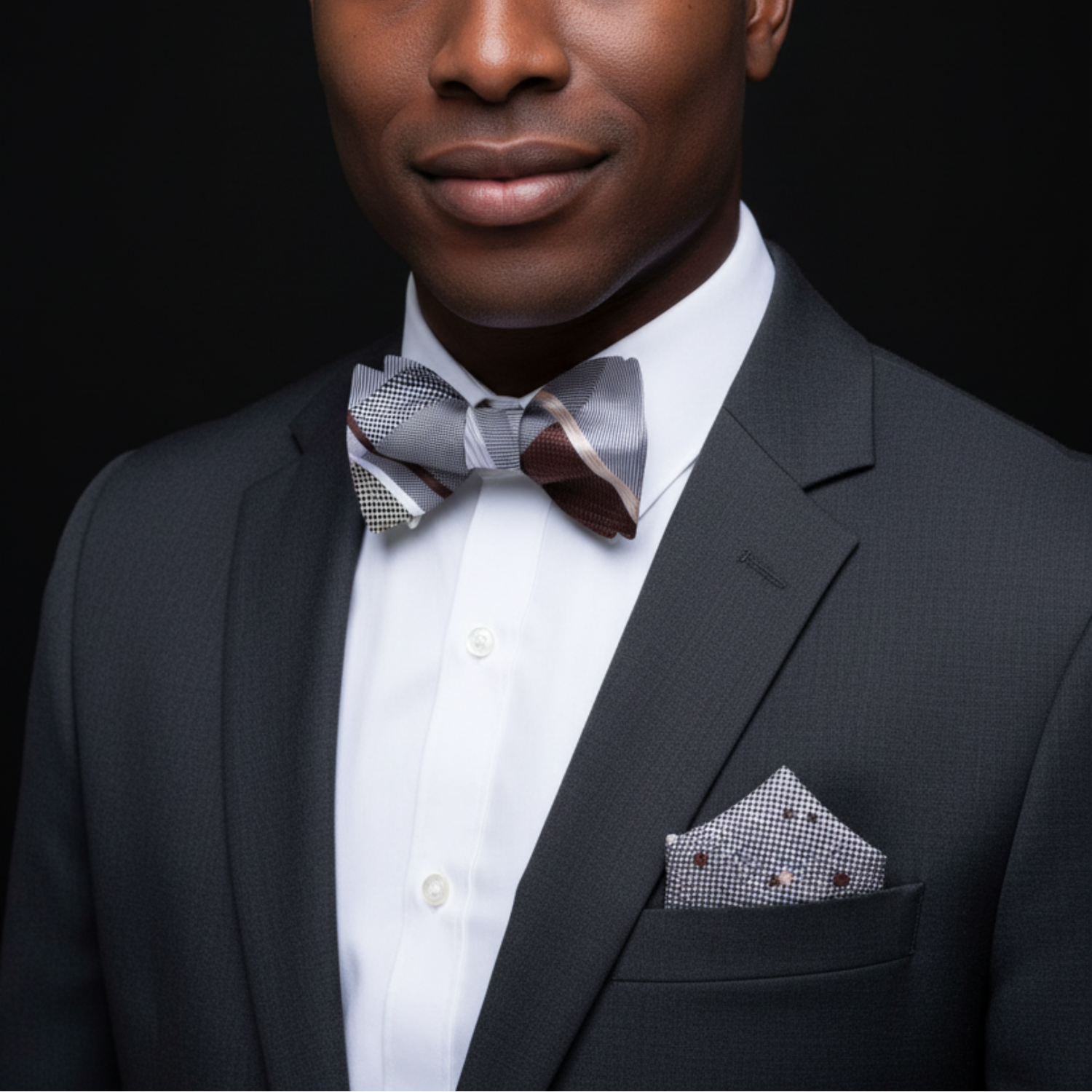 Man wearing a black suit with a white shirt, patterned bow tie, and pocket square against a dark background.
