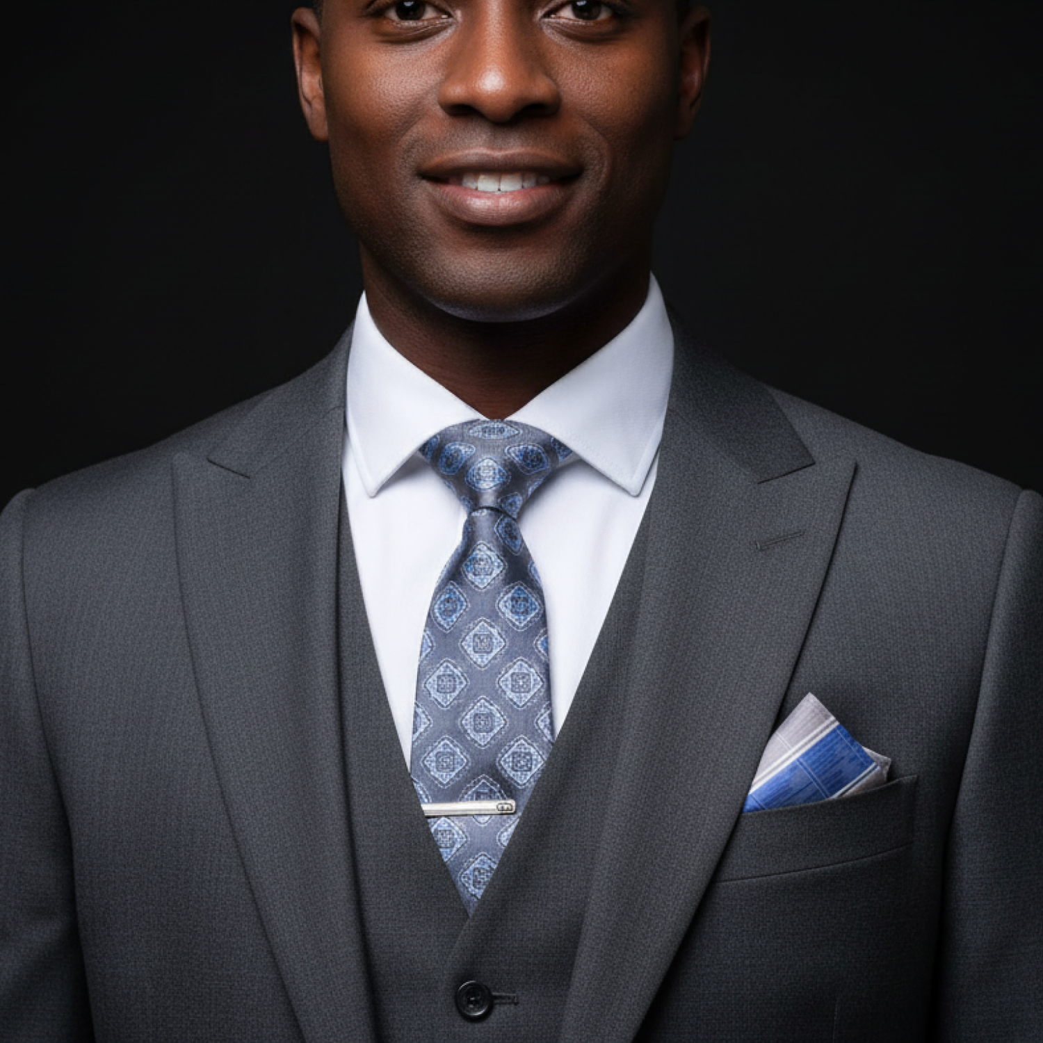 Man wearing a gray suit with a blue tie and accenting plaid square against a black background