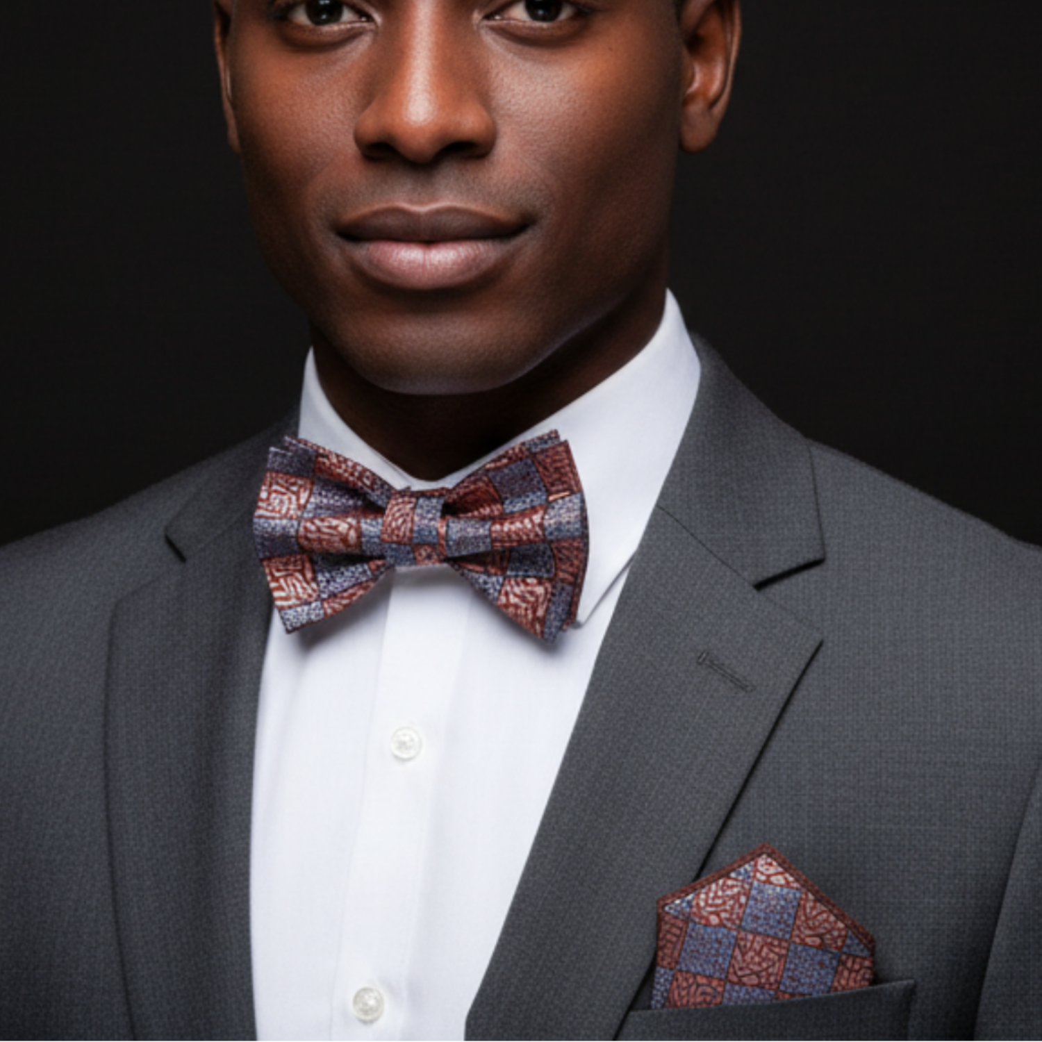 Man wearing a gray suit with a patterned bow tie and pocket square against a black background