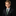 A young boy with styled light brown hair, wearing a dark navy suit, white dress shirt, and a textured green tie with a matching green pocket square, set against a solid black background.