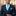 Man wearing a navy suit with a turquoise tie and pocket square, standing against a blurred cityscape background.
