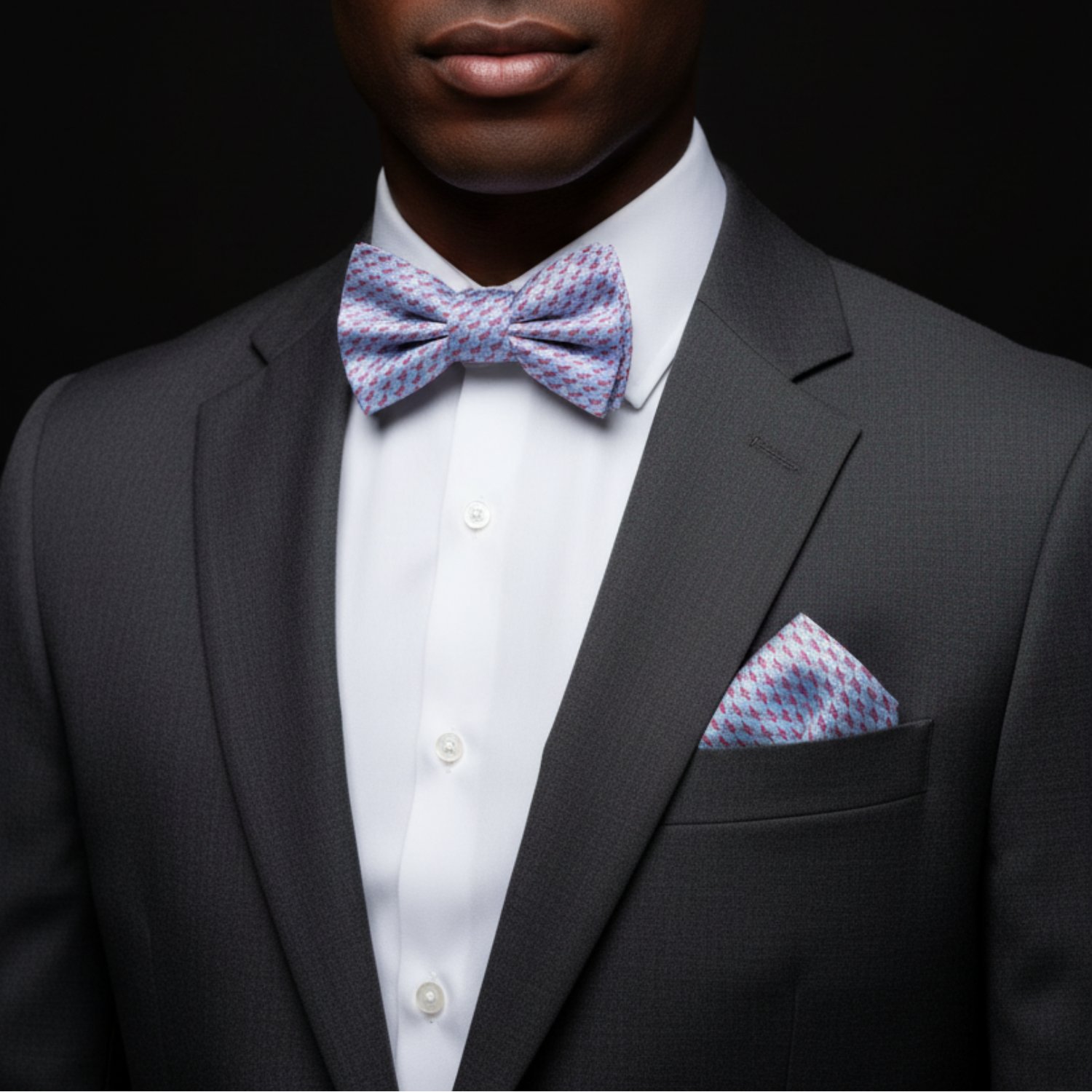 Man wearing a gray suit with a white shirt, patterned bow tie, and pocket square on a black background