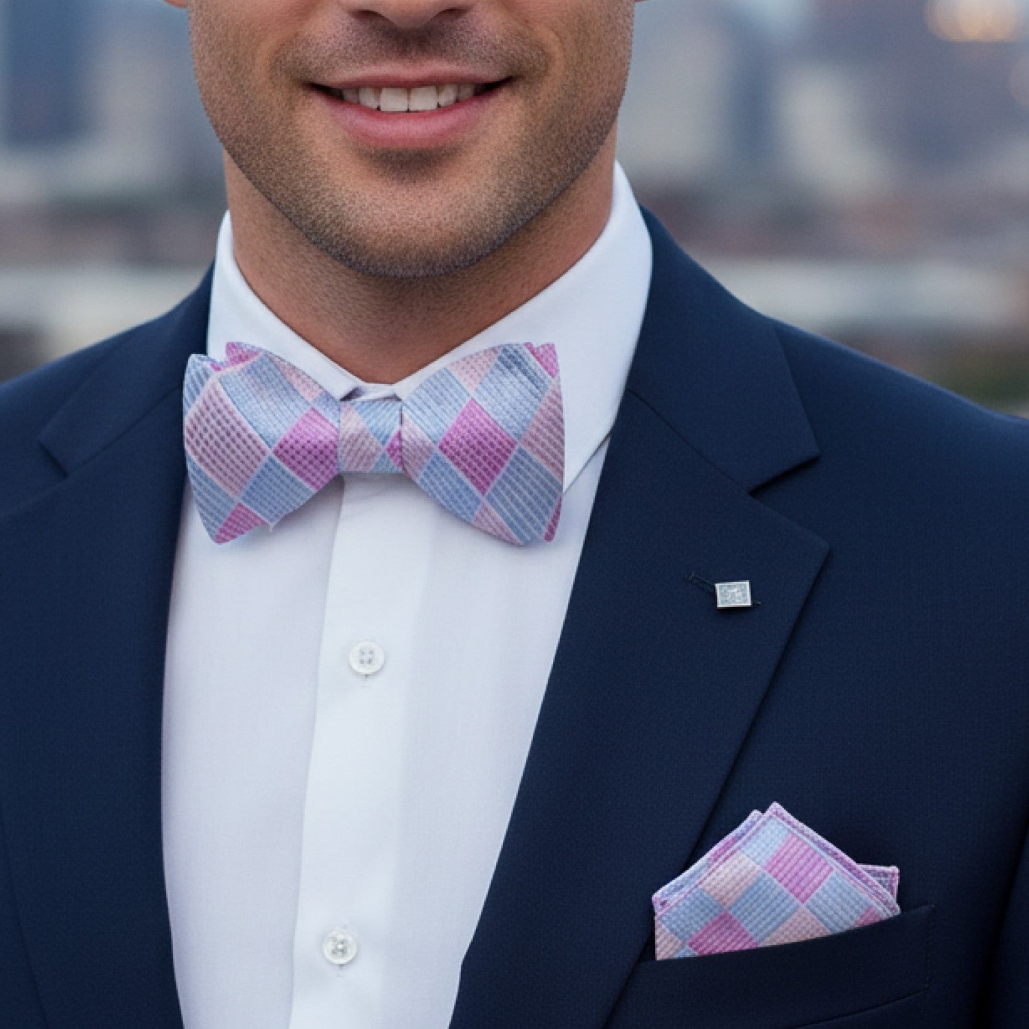 Man wearing a navy suit with a pink and blue checkered bow tie and pocket square, blurred cityscape in the background.
