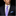 Man wearing a dark suit with a white shirt and blue patterned tie against a black background