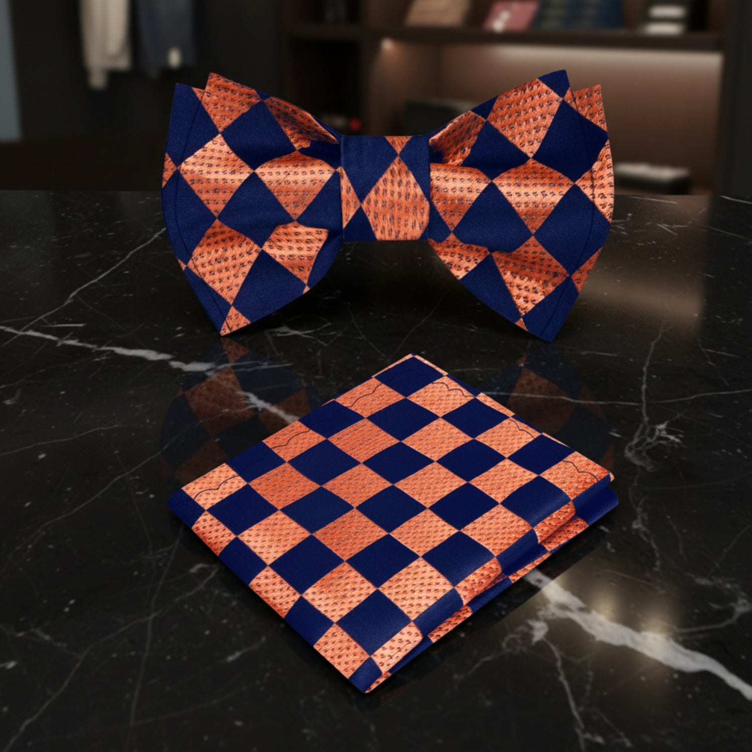 A matching men's formal accessory set featuring a pre-tied bow tie and a folded pocket square, both in a contrasting orange and navy blue checkerboard pattern with a subtle dotted texture.