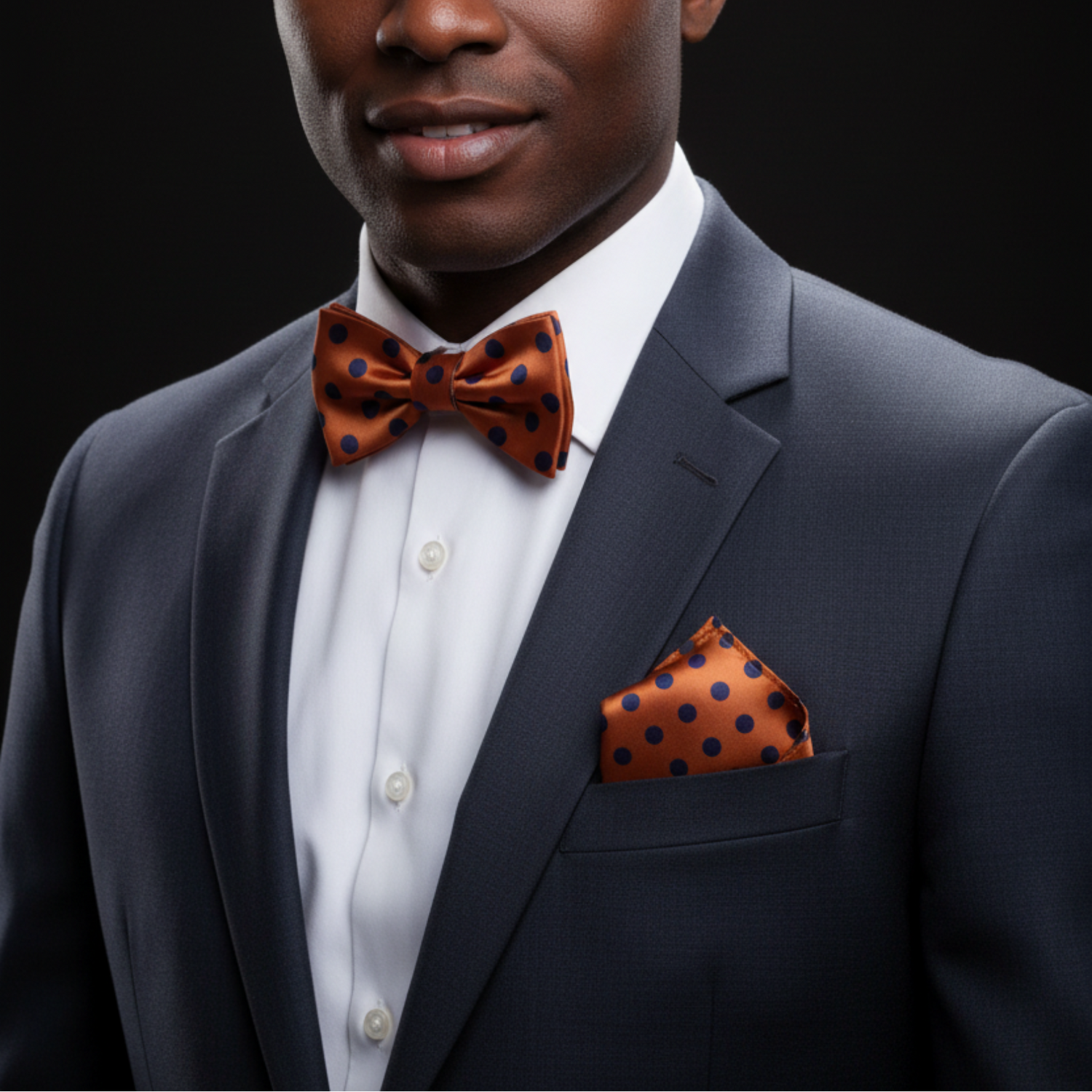 Man wearing a gray suit with an orange polka dot bow tie and pocket square on a black background