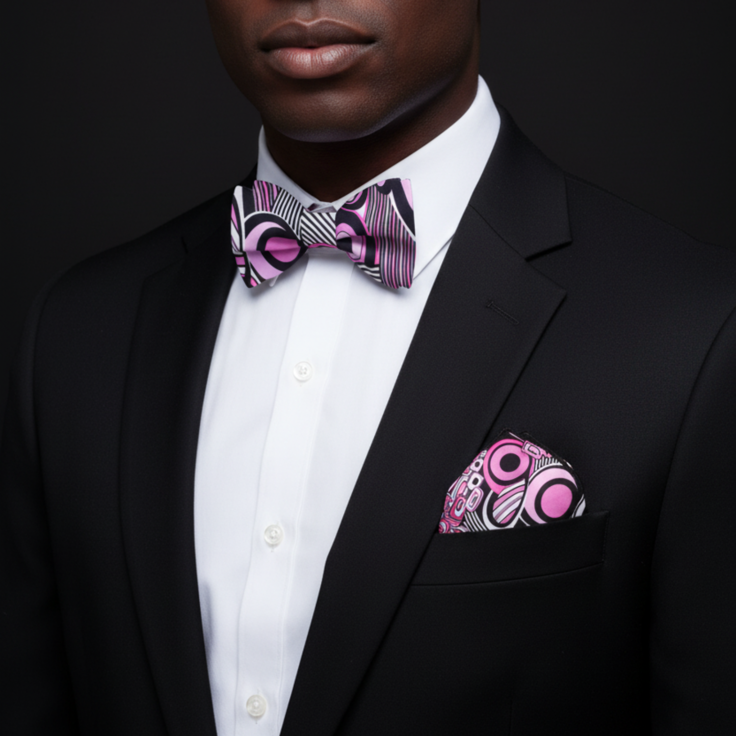 Man wearing a black suit with a white shirt, pink and purple patterned bow tie, and pocket square against a dark background.