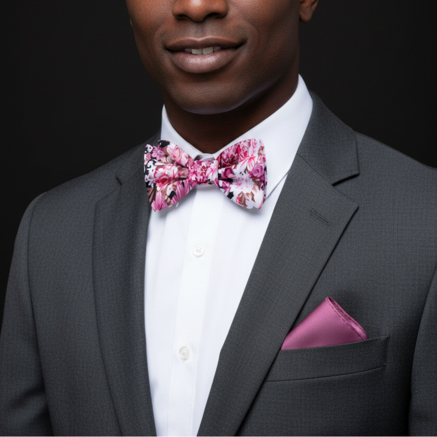 Man wearing a gray suit with a floral pink bow tie and matching pocket square on a black background