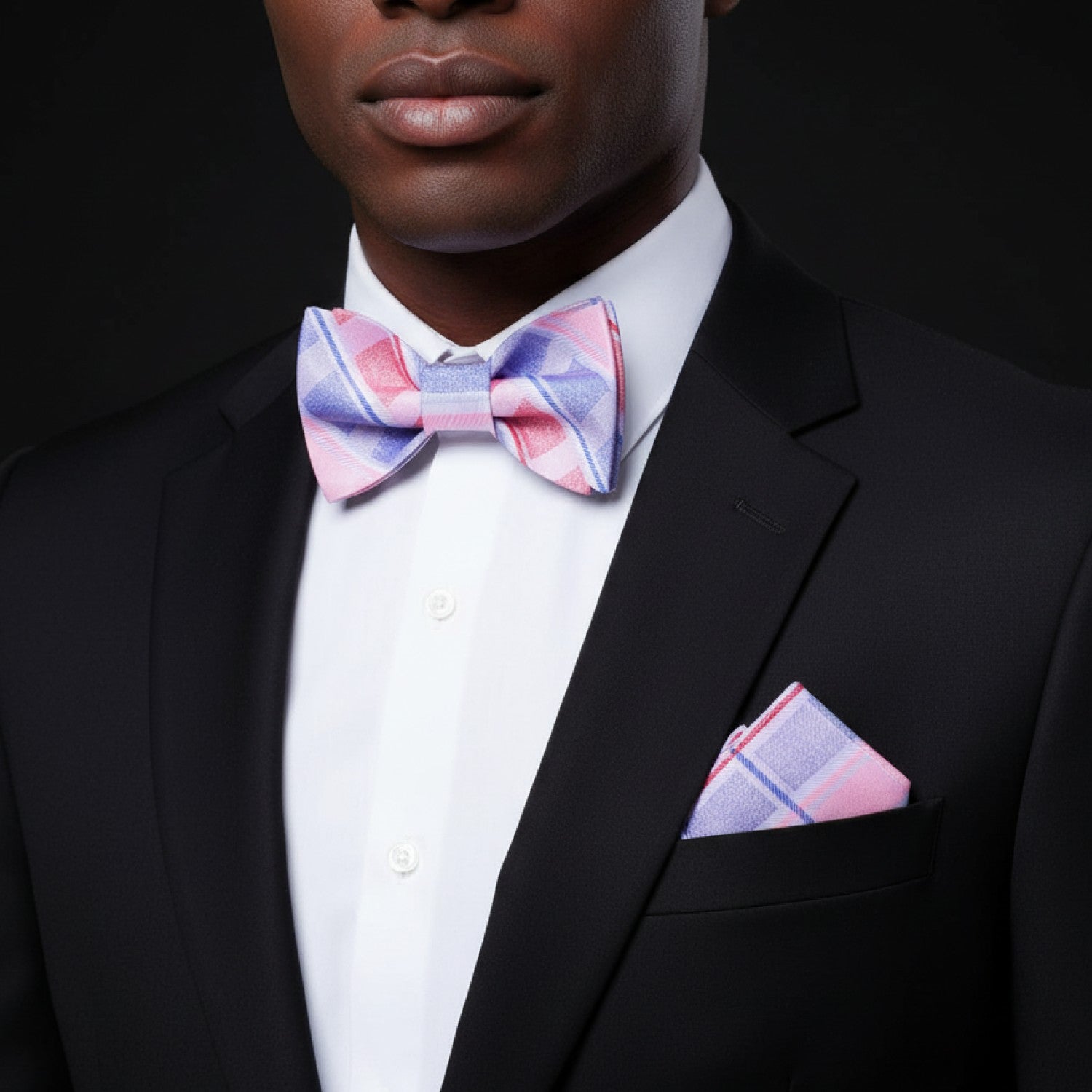 Man wearing a black suit with a pink and blue checkered bow tie and pocket square against a black background