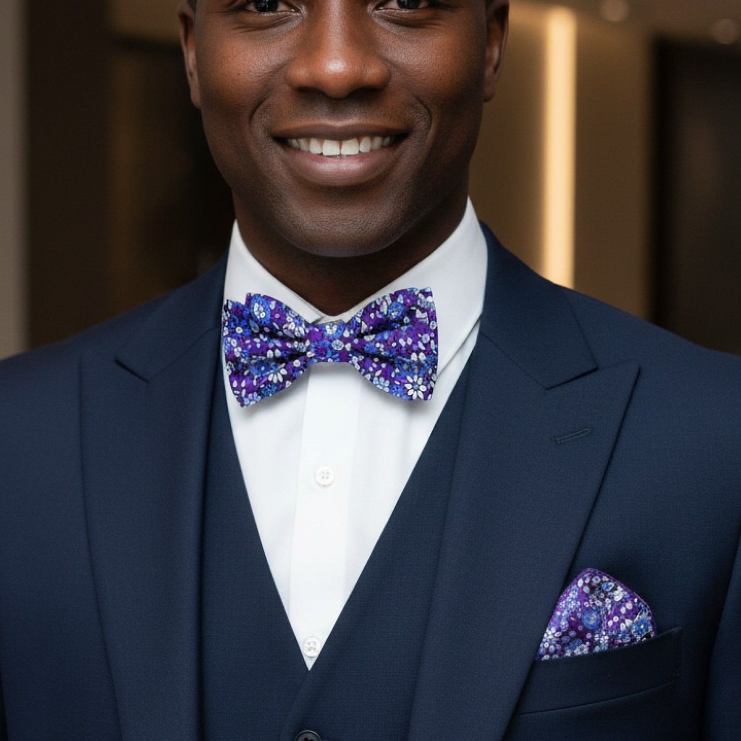 Man wearing a navy suit with a purple bow tie and pocket square, smiling.