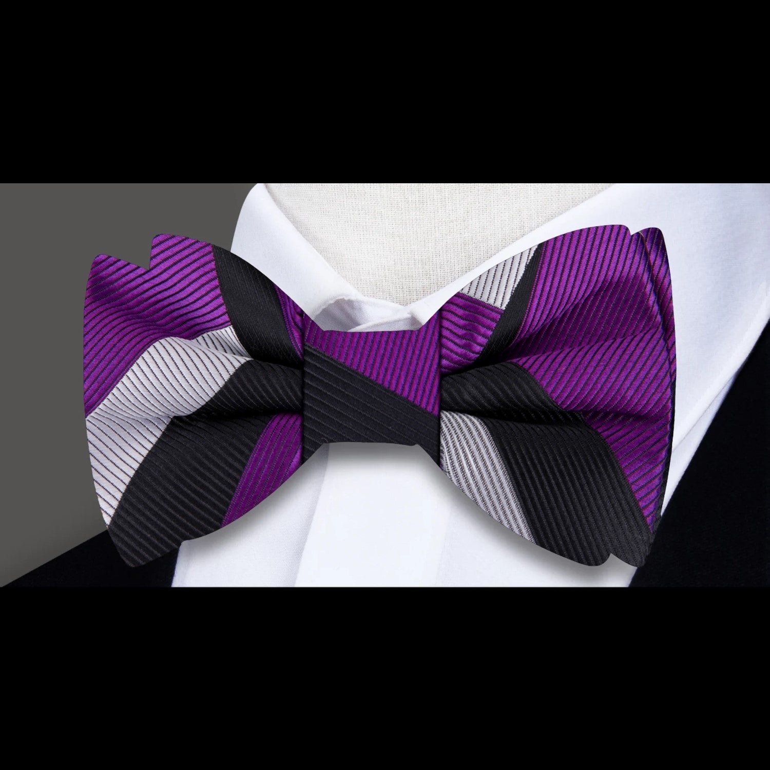 A pre-tied bow tie featuring geometric patterns in purple, black, and silver colors, displayed on a mannequin wearing a white shirt and black suit.