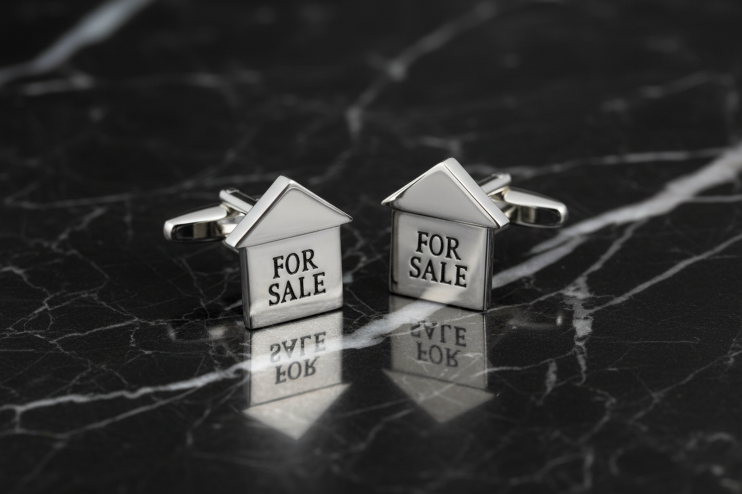 A pair of silver chrome cufflinks designed to resemble houses with 'FOR SALE' text on top, displayed on a red cushion.