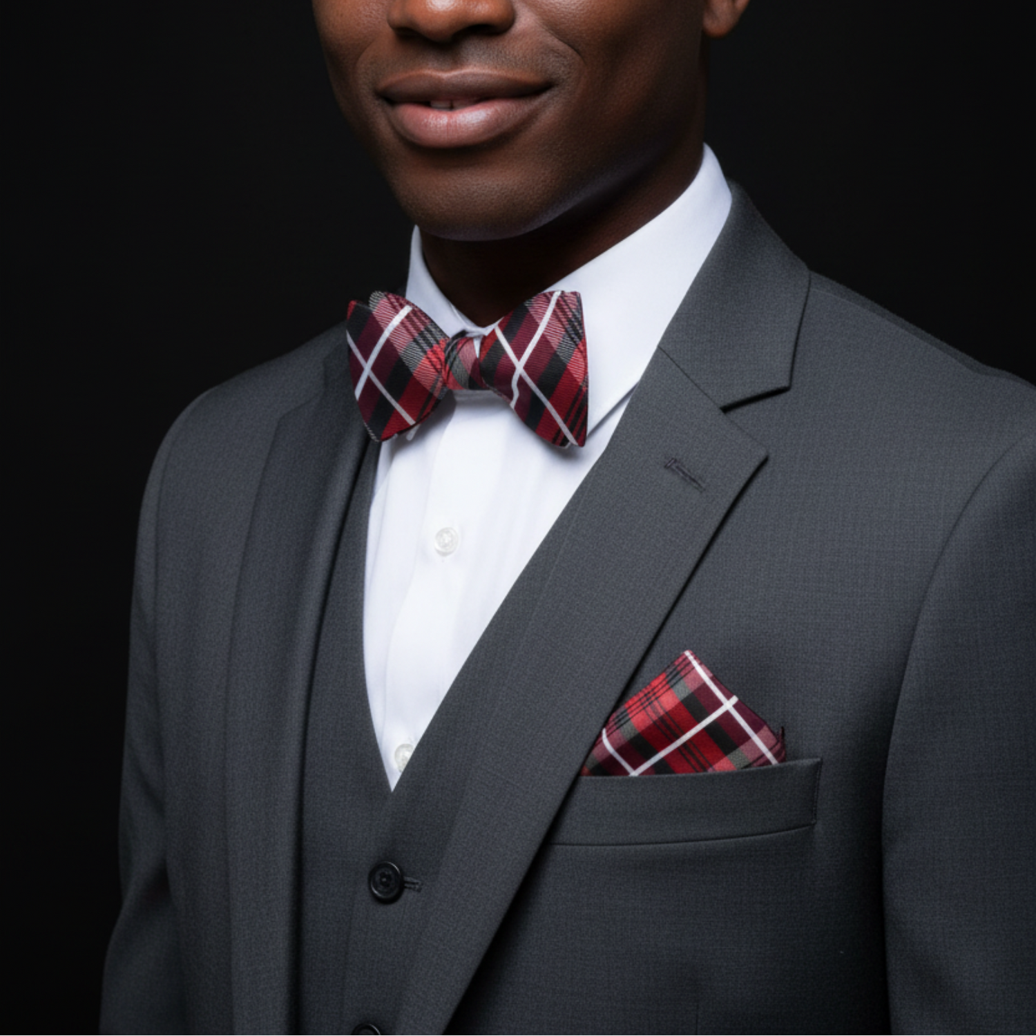Man wearing a gray suit with a red plaid bow tie and pocket square on a black background