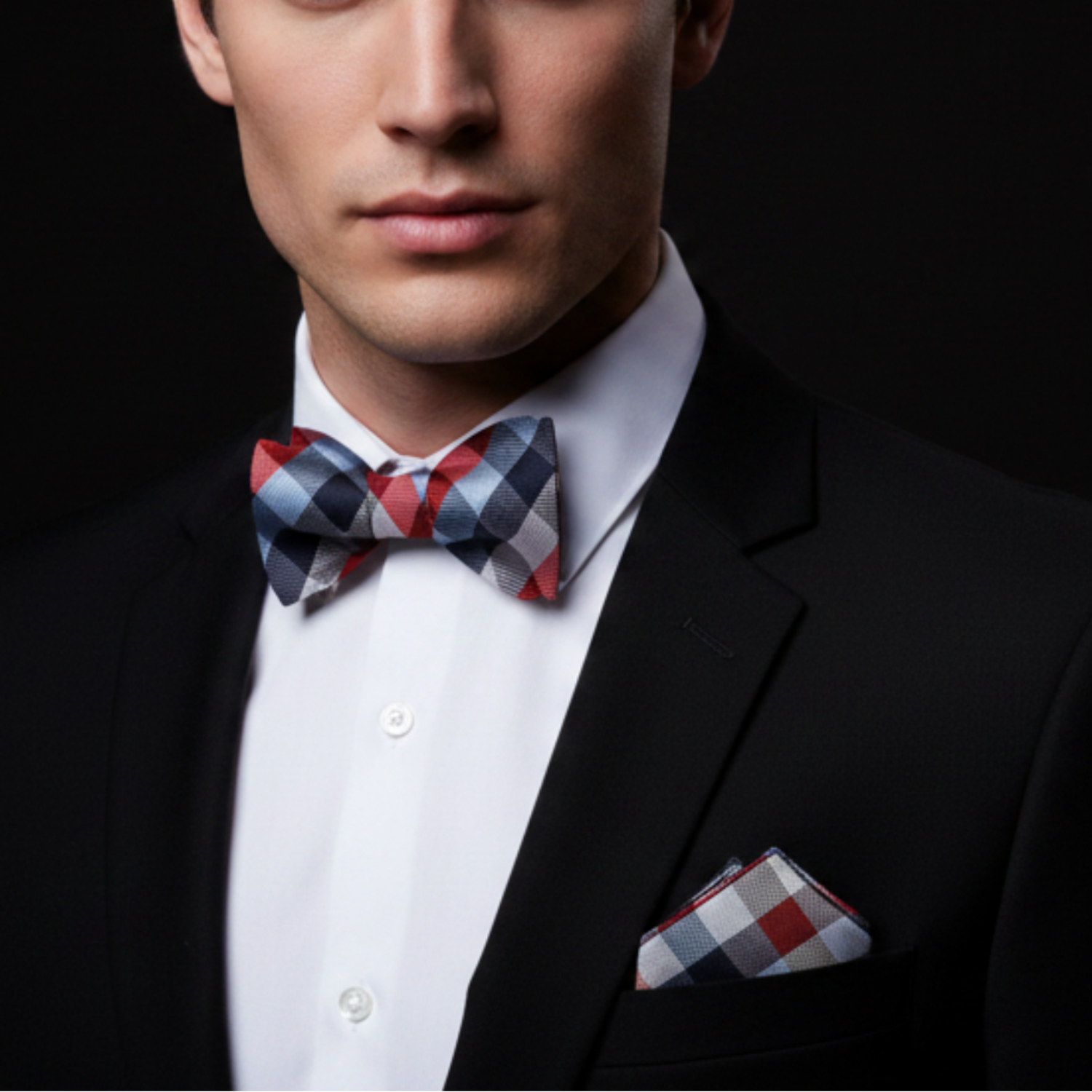 Man wearing a black suit with a plaid bow tie and pocket square on a dark background